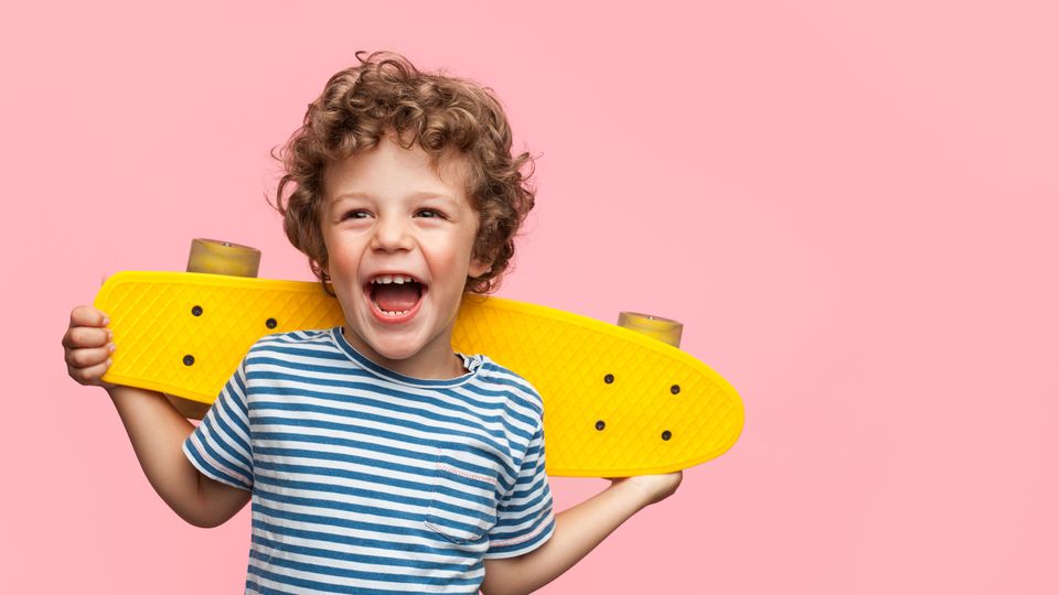 Kind Junge Skateboard Ein Junge mit braunen Locken und geringeltem T-Shirt lacht und hält dabei ein gelbes Skateboard hinter dem Rücken