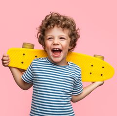 Ein Junge mit braunen Locken und geringeltem T-Shirt lacht und hält dabei ein gelbes Skateboard hinter dem Rücken