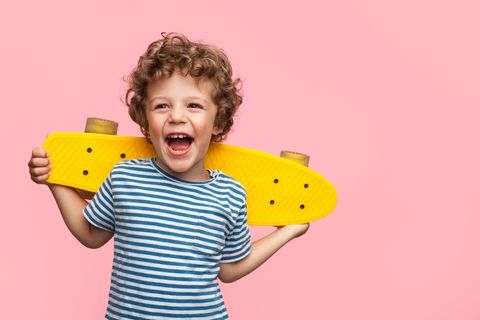 Ein Junge mit braunen Locken und geringeltem T-Shirt lacht und hält dabei ein gelbes Skateboard hinter dem Rücken