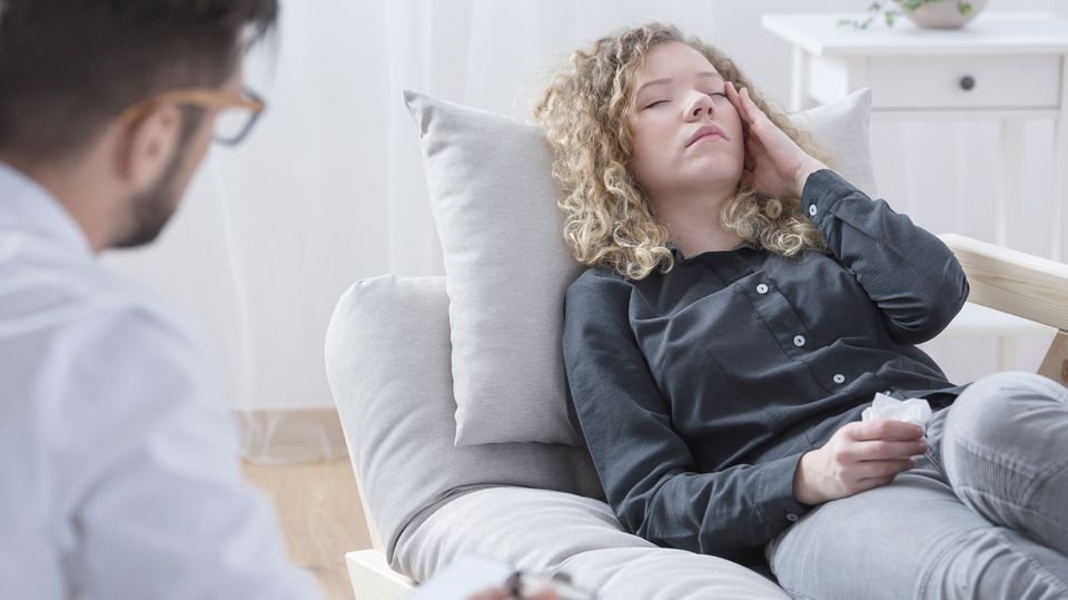 Eine Frau liegt auf einem Sofa und fässt sich mit einer Hand an den Kopf. Ein Mann sitzt ihr mit Zettel und Stift gegenüber