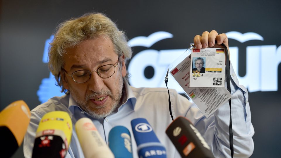 Can Dündar Der Journalist Can Dündar sitzt hinter vielen Mikrofonen und hält seine Akkreditierung für den Erdogan-Besuch mit links hoch
