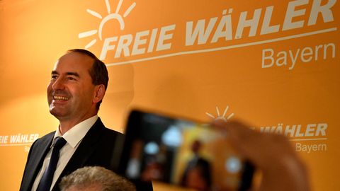 Vor einer orangen Wand mit weißem Schriftzug "Freie Wähler Bayern" steht ein Mann im Anzug und lächelt breit