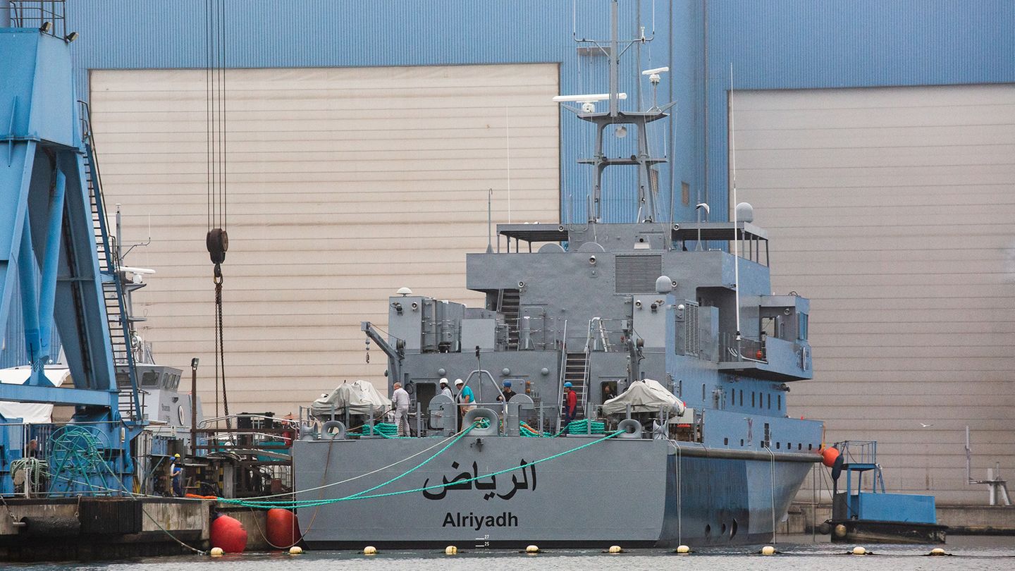Ein marinegraues Schiff liegt vor einer blauen Werfthalle mit weißen Rolltoren. Am Heck ein arabischer Schriftzug