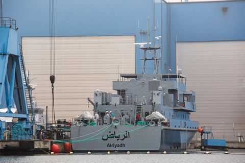 Ein marinegraues Schiff liegt vor einer blauen Werfthalle mit weißen Rolltoren. Am Heck ein arabischer Schriftzug