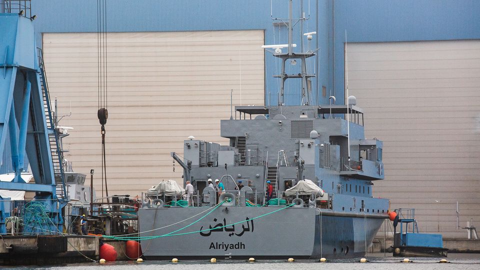 Ein marinegraues Schiff liegt vor einer blauen Werfthalle mit weißen Rolltoren. Am Heck ein arabischer Schriftzug