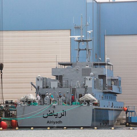 Ein marinegraues Schiff liegt vor einer blauen Werfthalle mit weißen Rolltoren. Am Heck ein arabischer Schriftzug