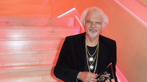 Ingo Interburg, ein Mann mit weißen Haaren und weißem Bart, steht bei einer Preisverleihung mit Trophäe vor einer roten Treppe