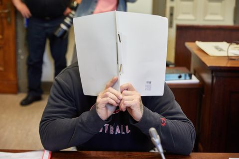 Ein Mann mit tätowierten Händen hält sich im Gerichtssaal einen weißen Schnellhefter vor sein Gesicht