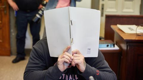 Ein Mann mit tätowierten Händen hält sich im Gerichtssaal einen weißen Schnellhefter vor sein Gesicht