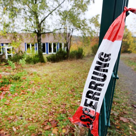An einem grünen Drahtzaun hängt ein Ende eines rot-weißen Flatterbandes mit "Polizeiabsperrung" darauf