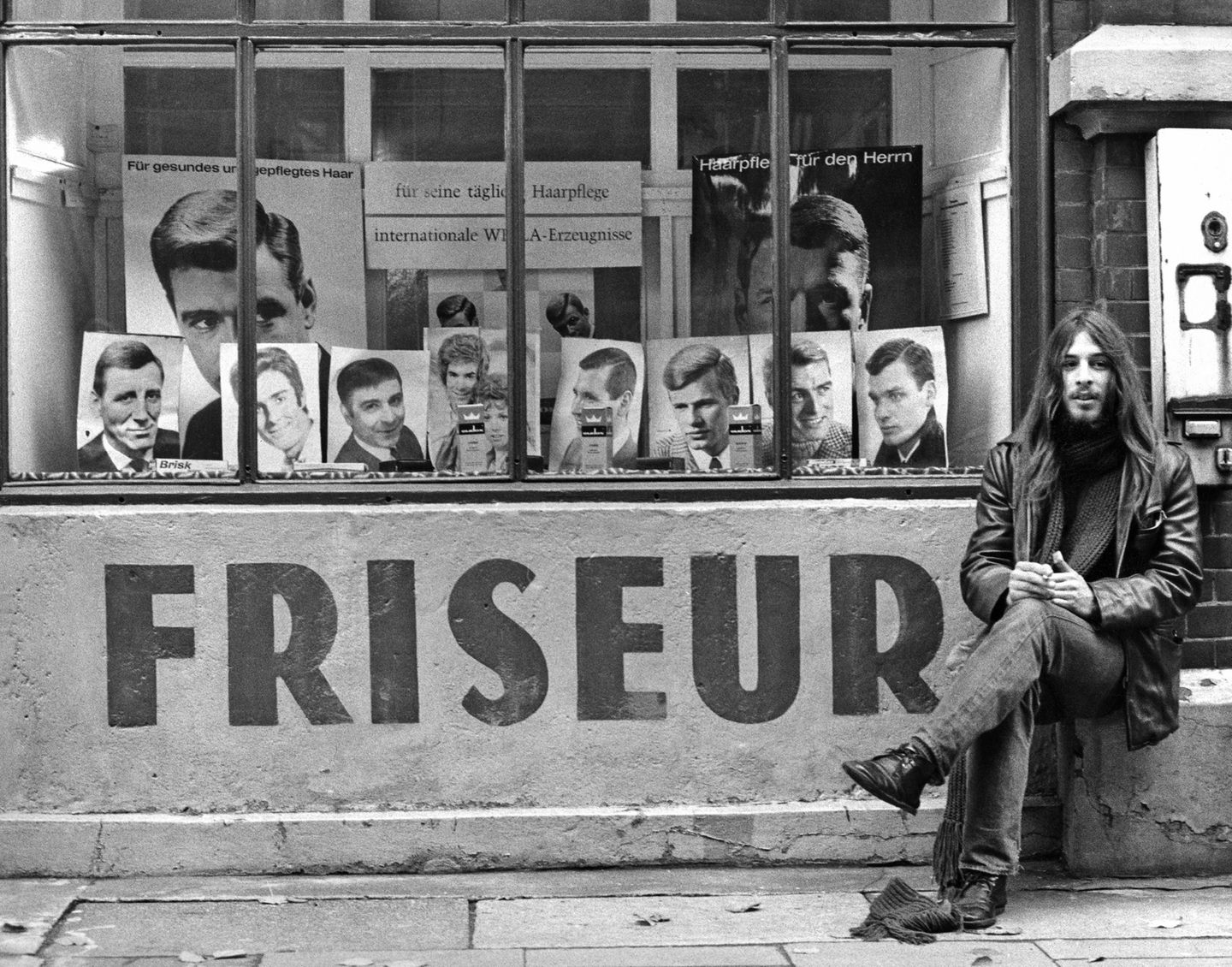 Ein langhaariger junger Mann vor einem Friseur-Schaufenster mit adretten Kurzhaarfrisuren