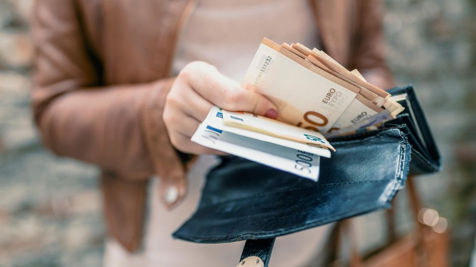 Eine Frau in hellbrauner Lederjacke zieht etliche Euroscheine aus ihrem großen Portemonnaie