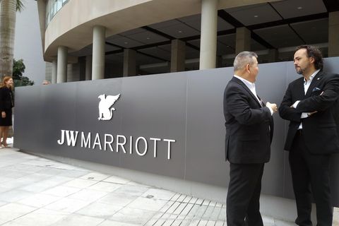Auf einer silbergrauen Metallwand ist der weiße Schriftzug "JW Marriott" zu sehen. Daneben stehen zwei Männer in Anzügen