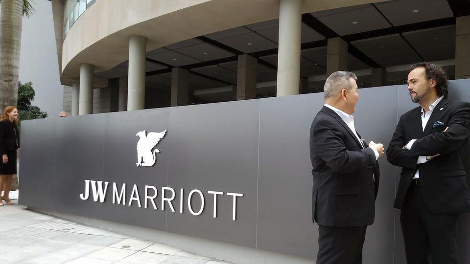 Auf einer silbergrauen Metallwand ist der weiße Schriftzug "JW Marriott" zu sehen. Daneben stehen zwei Männer in Anzügen