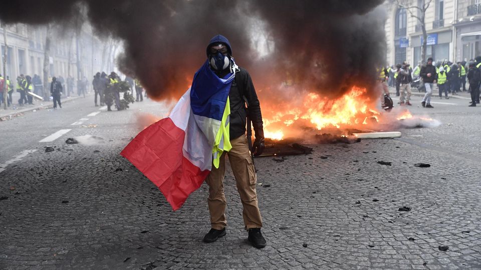 Paris, achtes Arrondissement: Ein "Gelbwesten"-Demonstrant posiert mit einer französischen Fahne vor einem brennenden Auto für die Kamera. Die Wut der Protestbewegung richtet sich aber auch gegen Präsident Macron und dessen Reformpolitik.