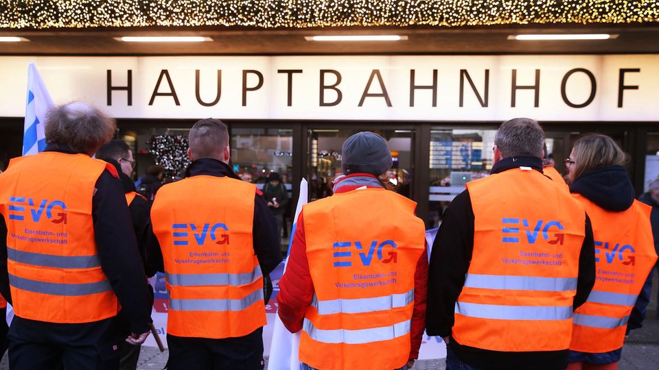 Fünf Männer in orangen Warnwesten mit EVG-Logo stehen vor einem "Hauptbahnhof"-Schild
