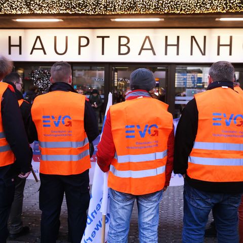 Fünf Männer in orangen Warnwesten mit EVG-Logo stehen vor einem "Hauptbahnhof"-Schild