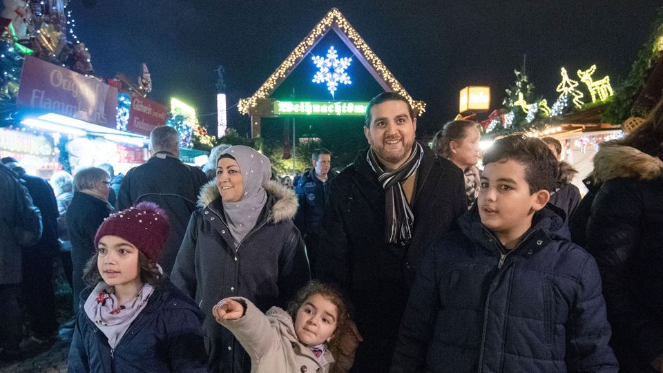 Eine fünfköpfige Familie geht über einen Weihnachtsmarkt. Die Augen der Kinder leuchten, ein kleines Mädchen zeigt auf etwas