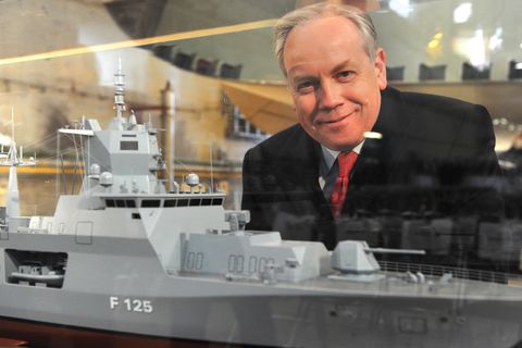 Ein Mann mit weißem Seitenscheitel lächelt durch einen Glaskasten mit dem Modell einer Fregatte hindurch