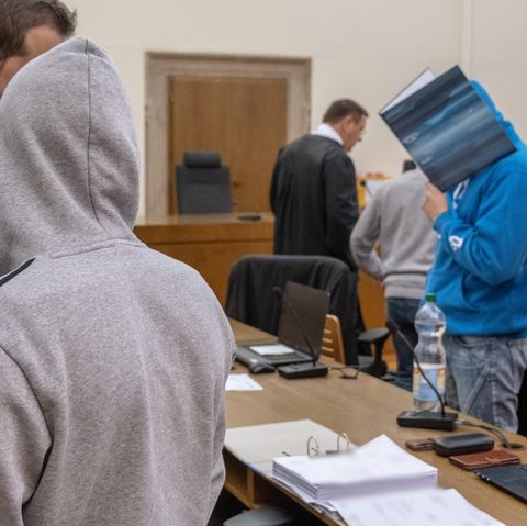 Die Angeklagten stehen im Gerichtssaal und verbergen sich unter den Kapuzen ihrer Pullover und hinter einem Aktenordner