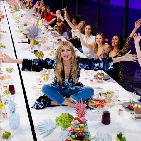 Heidi Klum sitzt auf einer langen Tafel und präsentiert die Kandidatinnen von GNTM 2019