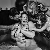 Nach einer Wassergeburt hält eine Mutter glücklich lachend ihr Baby im Arm, rund um das Wasserbecken freut sich die Familie