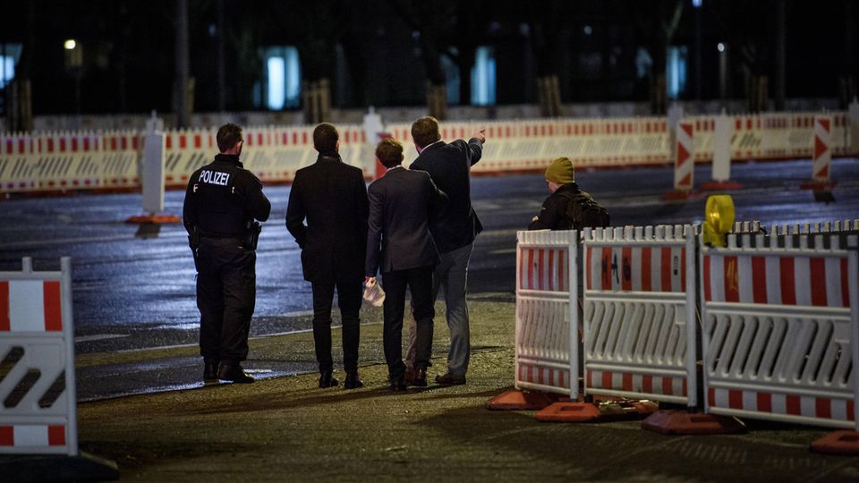 Im Dunkeln stehen drei junge Männer in Anzügen mit uniformierten Polizisten zwischen Baustellenabsperrungen. Einer zeigt etwas