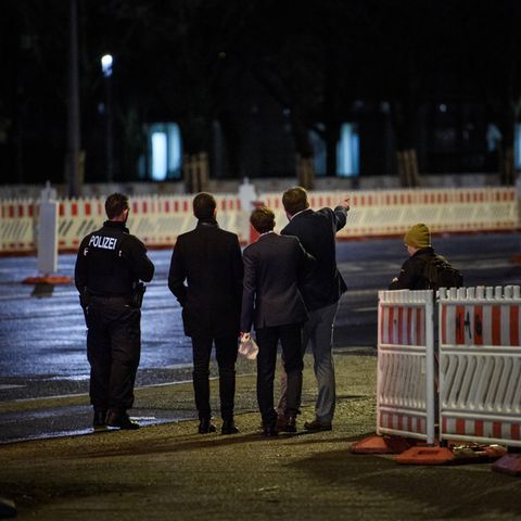 Im Dunkeln stehen drei junge Männer in Anzügen mit uniformierten Polizisten zwischen Baustellenabsperrungen. Einer zeigt etwas