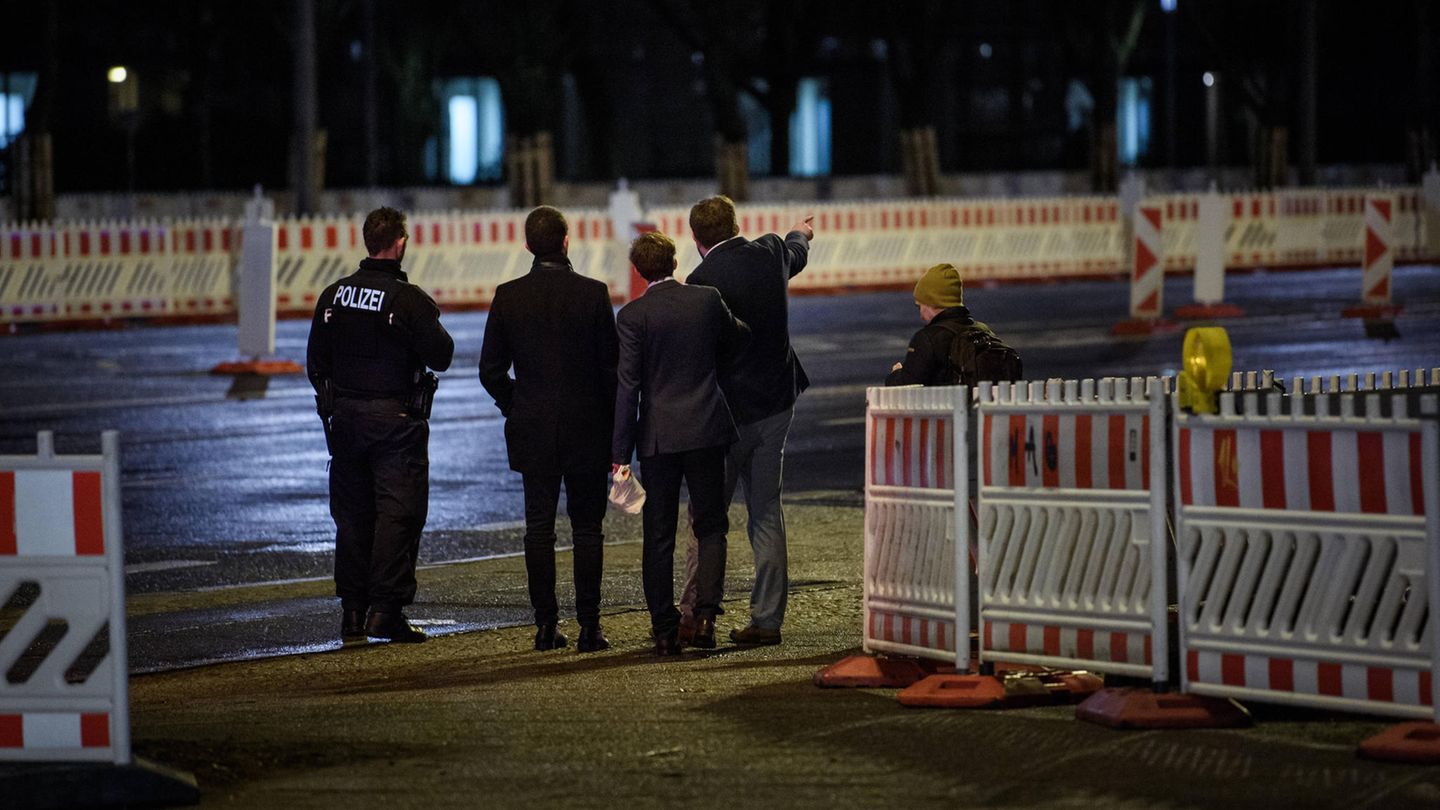 Im Dunkeln stehen drei junge Männer in Anzügen mit uniformierten Polizisten zwischen Baustellenabsperrungen. Einer zeigt etwas