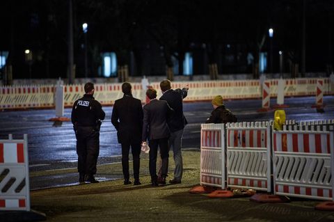 Im Dunkeln stehen drei junge Männer in Anzügen mit uniformierten Polizisten zwischen Baustellenabsperrungen. Einer zeigt etwas