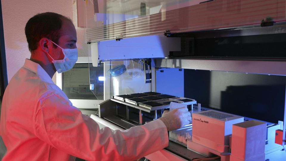 Ein Wissenschaftler mit Kittel und Mundschutz sitzt in einem Labor und hält Proben in seiner rechten Hand