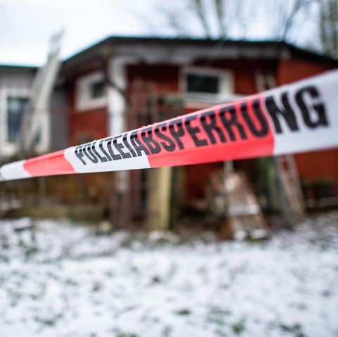 Vor einer rostroten Holzhütte hängt ein Absperrband "Polizeiabsperrung". Auf dem Rasen vor der Hütte liegt Schnee