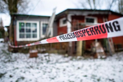 Vor einer rostroten Holzhütte hängt ein Absperrband "Polizeiabsperrung". Auf dem Rasen vor der Hütte liegt Schnee