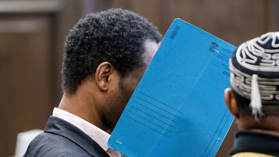 Ein Mann sitzt in einem Gerichtssaal und hält sich einen blauen Pappordner vors Gesicht