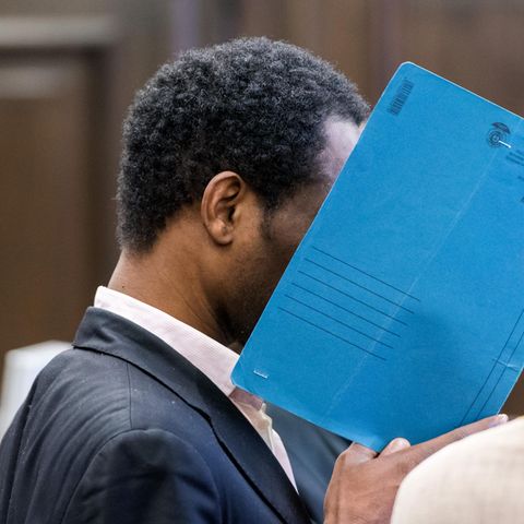 Ein Mann sitzt in einem Gerichtssaal und hält sich einen blauen Pappordner vors Gesicht