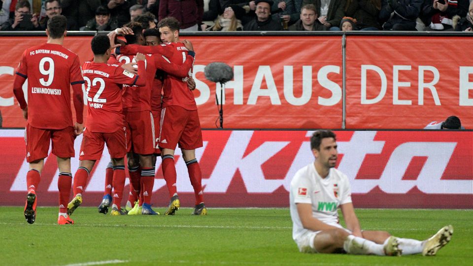 Augsburgs Rani Khedira (r.) sitzt niedergeschlagen auf dem Rasen, während die Bayern-Spieler jubeln