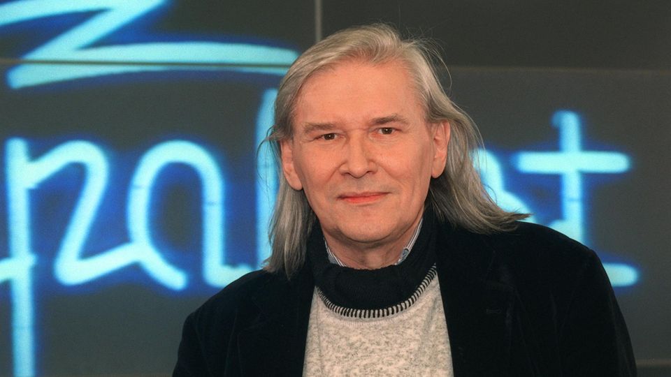 Vor einem Neon-Schriftzug" Rockpalast" schaut Peter Rüchel mit grauen, schulterlangen Haaren in die Kamera