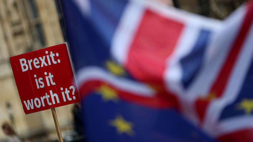 Eine EU- und eine Großbritannienflagge. Im Hintergrund ein Protestschild mit der Aufschrift "Brexit: is it worth it?"