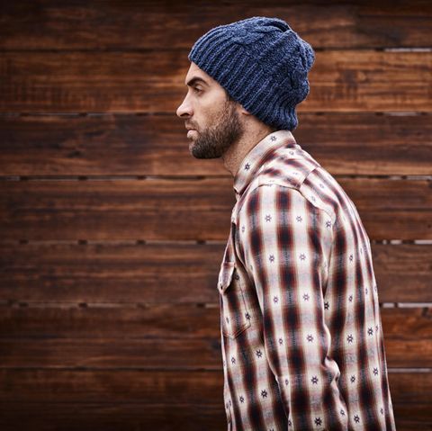 Ein junger Mann mit blauer Wollmütze und weiß-rot kariertem Hemd geht vor einer Bretterwand entlang