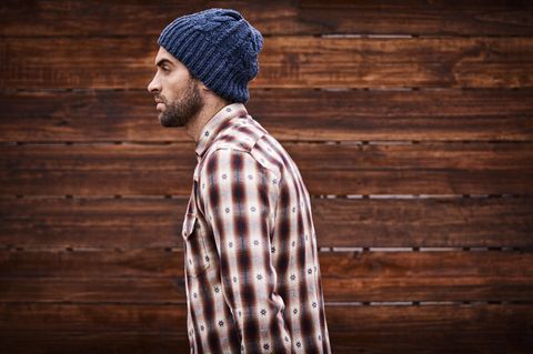 Ein junger Mann mit blauer Wollmütze und weiß-rot kariertem Hemd geht vor einer Bretterwand entlang