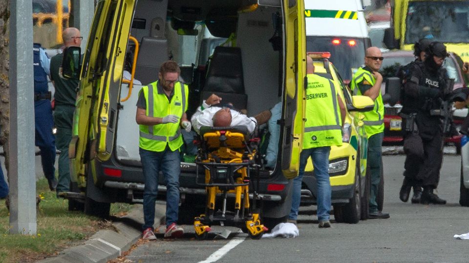Ein Verletzter wird in Christchurch in einen Krankenwagen geladen, während schwer bewaffnete Polizisten vorbeigehen
