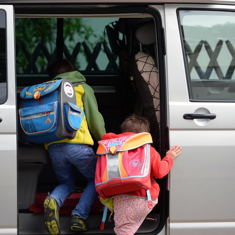 Zwei Grundschulkinder mit Ranzen auf dem Rücken steigen in die Schiebetür eines silbernen VW-Busses