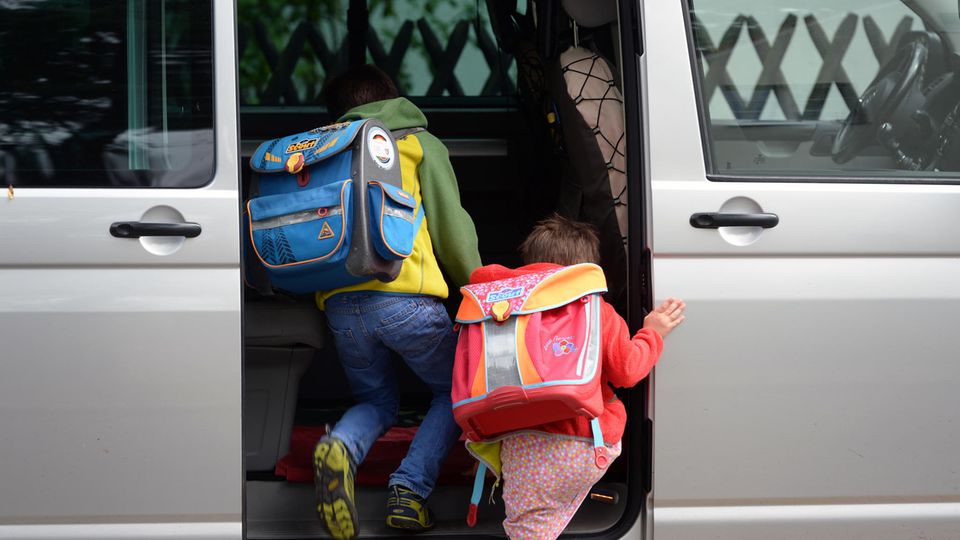 Zwei Grundschulkinder mit Ranzen auf dem Rücken steigen in die Schiebetür eines silbernen VW-Busses