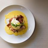 Eggs Benedict      Zutaten:  Brot-Muffins oder Pita-Brote Schinken Ei  Sauce hollandaise:  100 g ungesalzene Butter  2 Bio-Eigelbe  1 TL Senf  1 EL Weißweinessig  1 El Zitronensaft      Zubereitung:      Zuerst das Ei pochieren: Ei in einem Glas aufschlagen. Einen großen Topf mit Wasser zum Kochen bringen, Essig hinzufügen. Das siedende Wasser kreisförmig in Bewegung setzen und in die Mitte langsam das Ei hineingleiten lassen. Etwa zwei bis drei Minuten pochieren lassen. Dann das pochierte Ei aus dem Wasser schöpfen und kurz abtrocknen lassen.   Für die Sauce hollandaise Butter in einem Topf schmelzen lassen, über einem Wasserbad die Eigelbe in eine Schüssel geben und mit dem Zitronensaft und dem Senf schaumig schlagen. Dann langsam die Butter unterrühren. (Tipp: Falls die Sauce flockt, helfen ein paar Spritzer Wasser). Mit Essig abschmecken.  Schinken, Ei und Sauce hollandaise auf dem Pita-Brot anrichten und servieren.