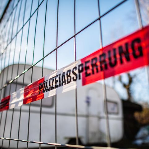 An einem Bauzaun vor einem Campingwagen hängt rot-weißes Band mit schwarzem Aufdruck "Polizeiabsperrung"