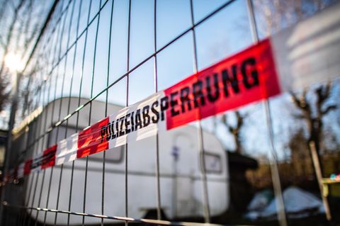 An einem Bauzaun vor einem Campingwagen hängt rot-weißes Band mit schwarzem Aufdruck "Polizeiabsperrung"