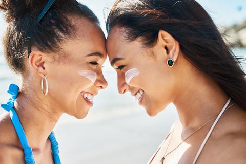 Zwei junge Frauen mit Sonnencreme auf den Wangen, stehen Stirn an Stirn und lächeln sich an