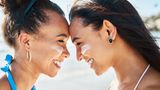 Zwei junge Frauen mit Sonnencreme auf den Wangen, stehen Stirn an Stirn und lächeln sich an