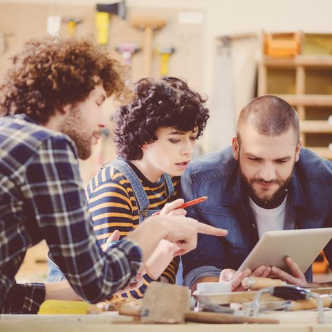 Zwei Handwerker und eine Handwerkerin besprechen etwas über Tablet gebeugt, in Werkstatt.