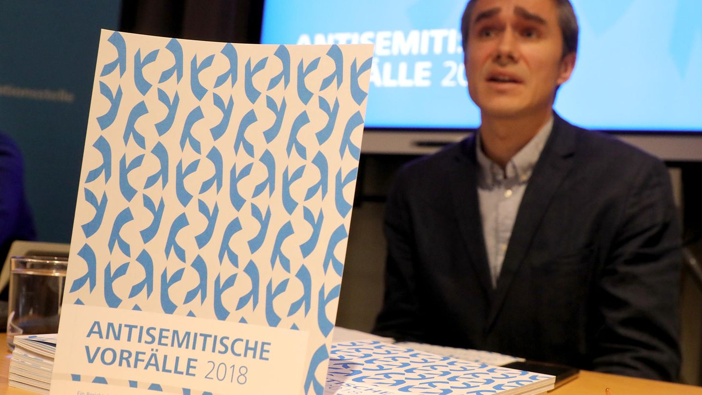 Eine weiß-blaue gemusterte Broschüre "Antisemitische Vorfälle 2018" steht auf einem Tisch. Dahinter sitzt ein Mann im Anzug